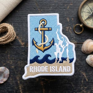 May include: Embroidered patch in the shape of Rhode Island, featuring a gold and navy anchor, waves, and the text "RHODE ISLAND". The patch has a light blue background and a white border. A nautical-themed item.