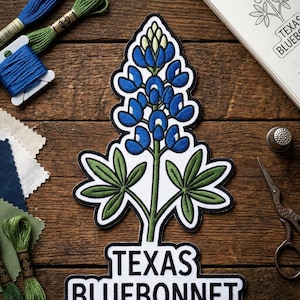 May include: An embroidered patch featuring a blue and green Texas Bluebonnet flower with a white background and black border. The text "TEXAS BLUEBONNET" is embroidered in black letters on a white rectangular base at the bottom of the patch.