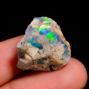 Peut inclure: Une pierre précieuse opale brute et non polie présentant une base blanc laiteux translucide avec des éclats vibrants de vert néon et de bleu électrique. La pierre a une forme irrégulière et dentelée avec une matrice rocheuse beige texturée.
