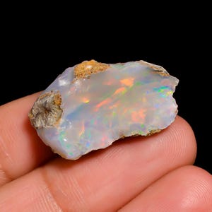 Op de afbeelding: Een ruwe, ongepolijste opaal edelsteen met een doorschijnende, melkwitte basis met levendige flitsen van iriserende oranje, groene en blauwe kleuren over het onregelmatige, grillige oppervlak en de textuurrijke bruine rotsranden.