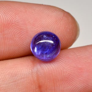 Puede incluir: Una gema redonda y pulida, de color azul profundo, posiblemente tanzanita, se sostiene entre dos dedos. La gema tiene una superficie lisa y reflectante con patrones internos sutiles y un reflejo brillante.