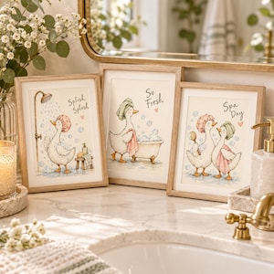 Op de afbeelding: Drie ingelijste wandkunstprints met aquarelillustraties van eenden in een badkamer. De eerste print toont een eend onder de douche met de tekst Splish Splash. De tweede toont een eend in een badkuip met So Fresh. De derde toont twee eenden met Spa Day.