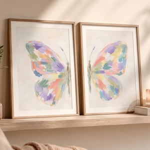 May include: Two framed wall art prints featuring a split butterfly design. Each print displays half of a butterfly with visible brushstrokes in pastel shades of purple, pink, yellow, green, and blue against a neutral, off-white background.