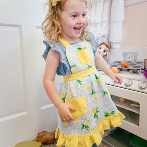 Puede incluir: Una ni&ntilde;a peque&ntilde;a con un delantal azul y blanco con estampado de limones y detalles amarillos, incluyendo un bolsillo y un dobladillo con volantes. La ni&ntilde;a tambi&eacute;n tiene un lazo amarillo en el pelo. El delantal es parte de un juego de cocina.