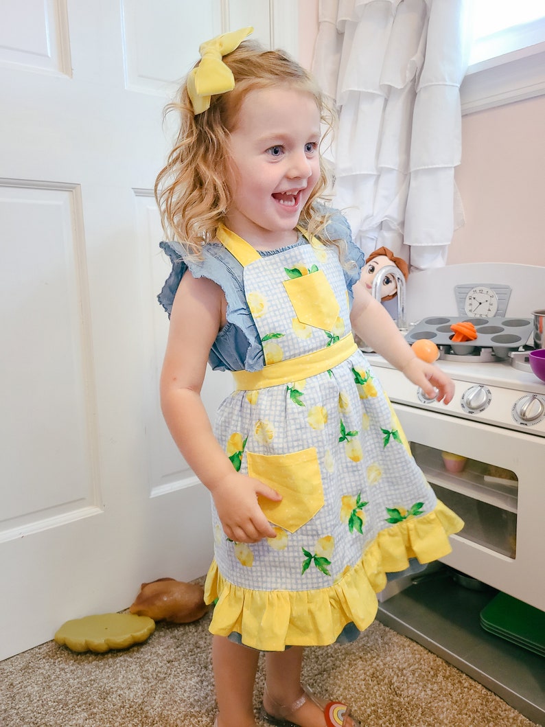 Puede incluir: Una ni&ntilde;a peque&ntilde;a con un delantal azul y blanco con estampado de limones y detalles amarillos, incluyendo un bolsillo y un dobladillo con volantes. La ni&ntilde;a tambi&eacute;n tiene un lazo amarillo en el pelo. El delantal es parte de un juego de cocina.