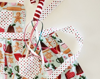 May include: A light blue apron with a festive Christmas baking theme. The apron features a pattern of gingerbread men, holly, and baking tools. A candy cane wand with red and white stripes and ribbons is in the center. The apron has a white polka dot trim.