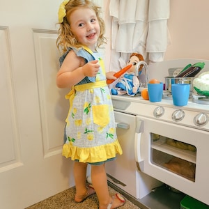 Puede incluir: Una ni&ntilde;a lleva un delantal azul y amarillo con estampado de limones y un lazo amarillo en el pelo. La ni&ntilde;a est&aacute; junto a una cocina de juguete con una mu&ntilde;eca y verduras de juguete.