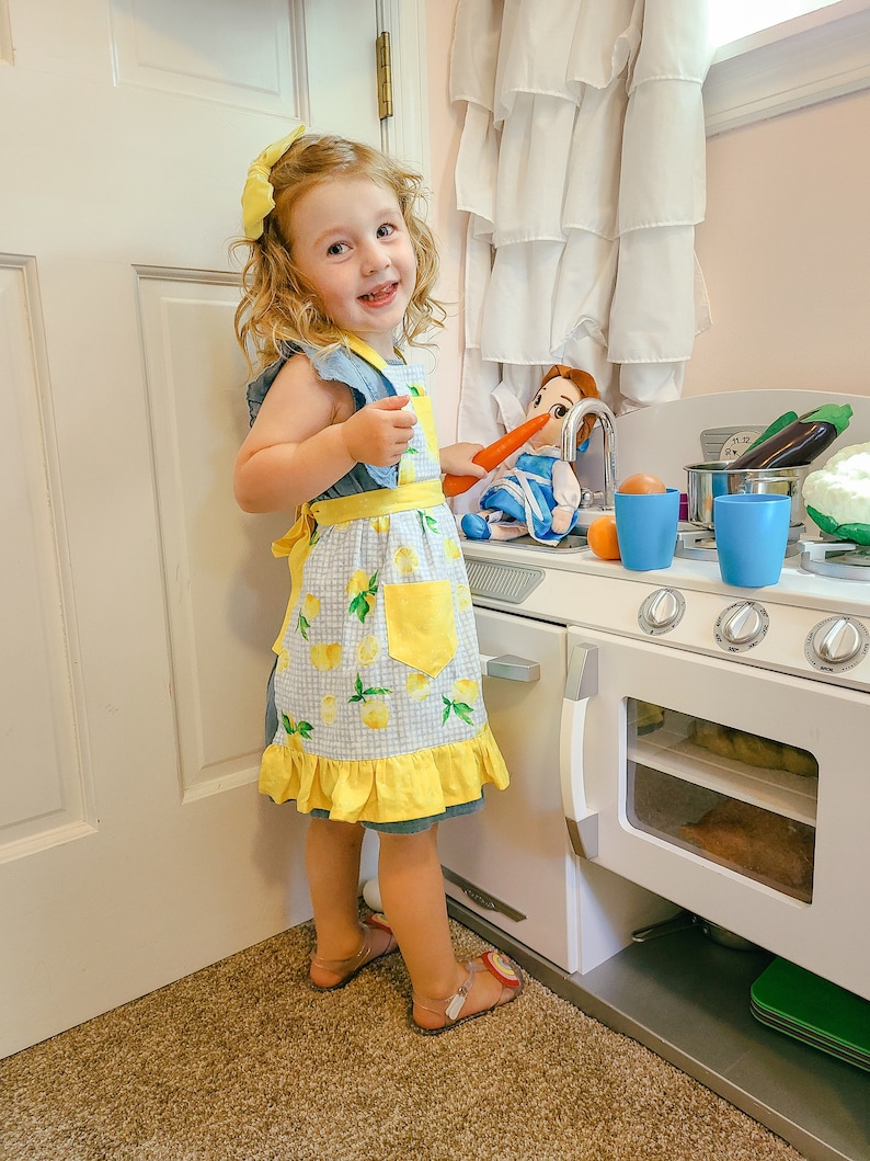 Puede incluir: Una ni&ntilde;a lleva un delantal azul y amarillo con estampado de limones y un lazo amarillo en el pelo. La ni&ntilde;a est&aacute; junto a una cocina de juguete con una mu&ntilde;eca y verduras de juguete.