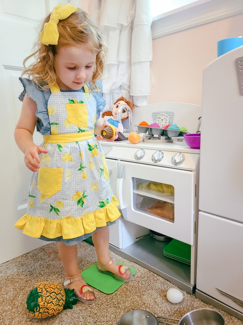 Puede incluir: Una ni&ntilde;a con un lazo amarillo y un delantal con estampado de limones est&aacute; de pie cerca de una cocina de juguete blanca. El delantal tiene un bolsillo y un ribete amarillos. Tambi&eacute;n se ven una pi&ntilde;a de juguete y una mu&ntilde;eca. La ni&ntilde;a lleva sandalias transparentes con detalles de arco&iacute;ris.