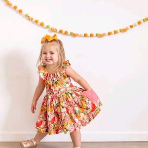 Può includere: Una bambina indossa un vestito rosa a fiori con un motivo di frutta, tra cui limoni, arance e angurie. L'abito ha maniche a balze e una tasca abbinata. Indossa sandali dorati e un grande fiocco arancione. Una ghirlanda di frutta in feltro decora lo sfondo.