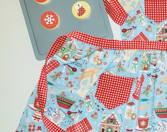 May include: A light blue Christmas apron with a red and white checkered trim and pocket. The apron features a festive pattern of gingerbread houses, snowmen, and cookies. A baking sheet with Christmas cookie designs is in the background.