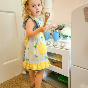 Puede incluir: Una ni&ntilde;a peque&ntilde;a con un lazo amarillo y un delantal azul y amarillo con tem&aacute;tica de limones, sosteniendo una esp&aacute;tula de madera. La ni&ntilde;a est&aacute; jugando con una cocina de juguete blanca, que incluye una estufa, ollas y verduras de juguete.