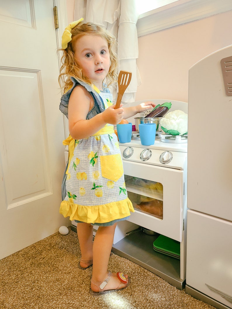 Puede incluir: Una ni&ntilde;a peque&ntilde;a con un lazo amarillo y un delantal azul y amarillo con tem&aacute;tica de limones, sosteniendo una esp&aacute;tula de madera. La ni&ntilde;a est&aacute; jugando con una cocina de juguete blanca, que incluye una estufa, ollas y verduras de juguete.