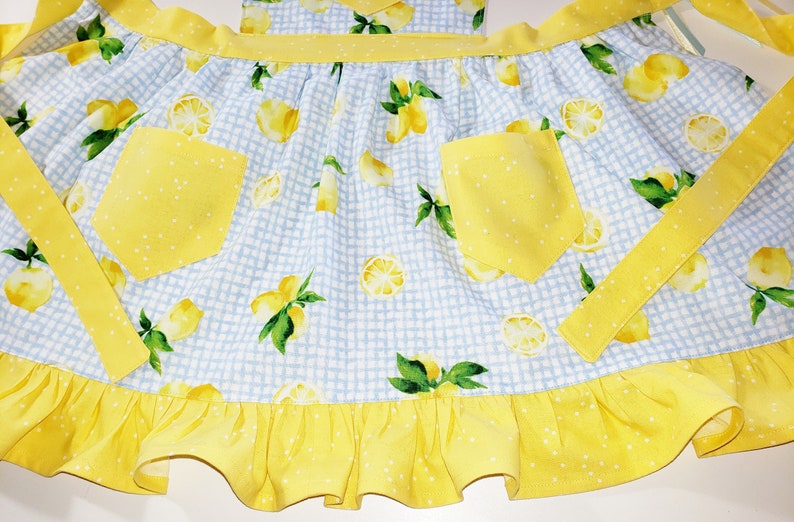 Puede incluir: Delantal a cuadros azules y blancos con un estampado de limones. El delantal tiene dos bolsillos amarillos con lunares y un dobladillo con volantes. Lazos y ribetes amarillos con lunares completan el dise&ntilde;o.
