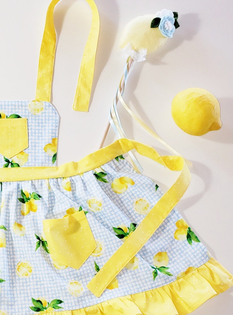 Puede incluir: Un delantal a cuadros azul y blanco con un bolsillo amarillo y estampado de lim&oacute;n. Una varita m&aacute;gica con forma de lim&oacute;n amarillo con cintas y un lim&oacute;n amarillo tambi&eacute;n est&aacute;n en la imagen. El delantal tiene correas y ribetes amarillos.