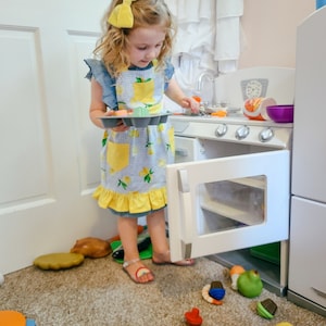 Puede incluir: Una ni&ntilde;a con un lazo amarillo y un delantal con estampado de limones juega con una cocina de juguete. La ni&ntilde;a sostiene una bandeja de cupcakes de juguete, con m&aacute;s comida de juguete esparcida por el suelo. La cocina de juguete es blanca con detalles plateados.