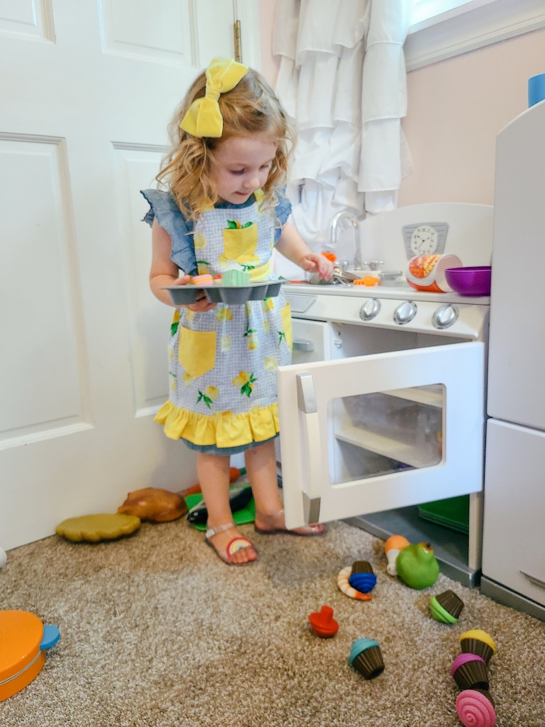 Puede incluir: Una ni&ntilde;a con un lazo amarillo y un delantal con estampado de limones juega con una cocina de juguete. La ni&ntilde;a sostiene una bandeja de cupcakes de juguete, con m&aacute;s comida de juguete esparcida por el suelo. La cocina de juguete es blanca con detalles plateados.