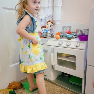 Puede incluir: Una ni&ntilde;a con un vestido de mezclilla y un delantal con estampado de limones, con un lazo amarillo en el pelo, est&aacute; junto a una cocina de juguete blanca. La cocina tiene una estufa, un horno y varios alimentos de juguete, incluyendo cupcakes y una olla.