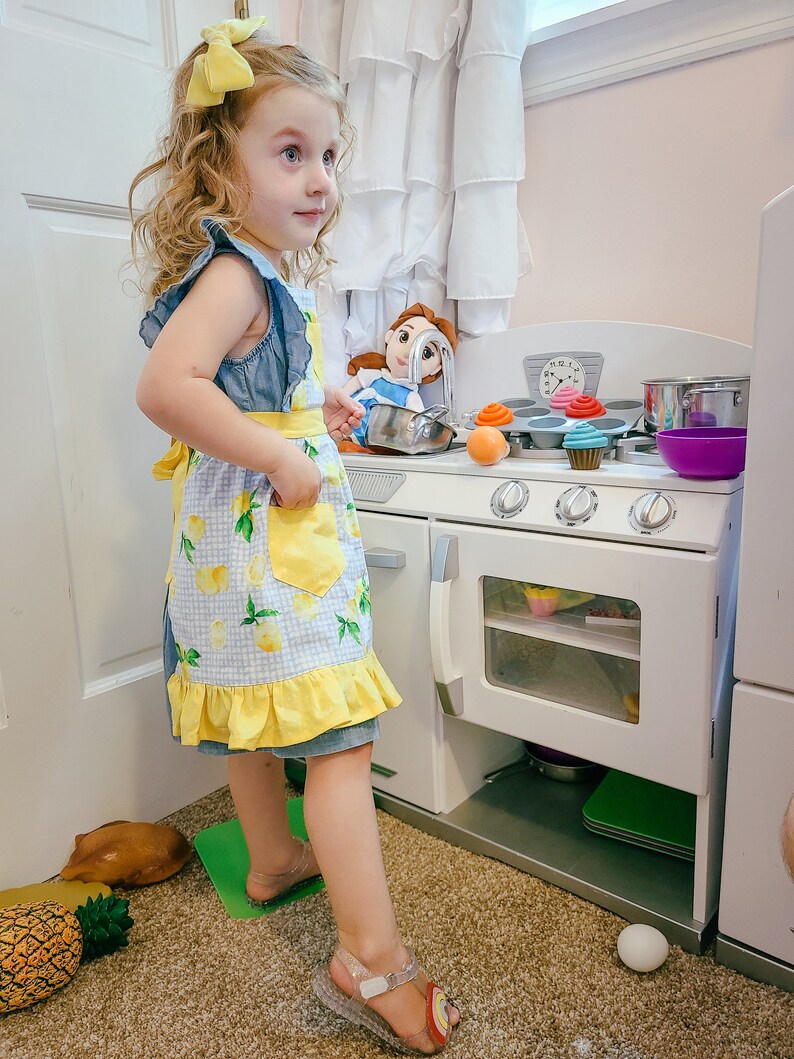 Puede incluir: Una ni&ntilde;a con un vestido de mezclilla y un delantal con estampado de limones, con un lazo amarillo en el pelo, est&aacute; junto a una cocina de juguete blanca. La cocina tiene una estufa, un horno y varios alimentos de juguete, incluyendo cupcakes y una olla.