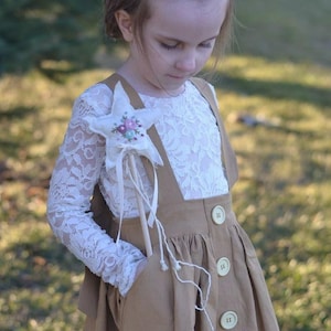 May include: A young girl wearing a white lace long-sleeved shirt and a brown overall skirt with buttons. She is holding a white star-shaped wand with a floral embroidery design.