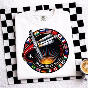 Puede incluir: Camiseta blanca con un diseño circular que muestra un cohete, banderas y el texto "PROJECT HAIL MARY". El diseño incluye un arco rojo y negro, una estrella y un sol radiante.