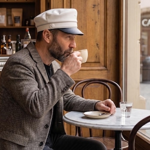 May include: A person wearing a white cap and a brown tweed jacket sips from a white coffee cup at a cafe. A small marble table holds a saucer, spoon, and a glass of water. The cafe has a wooden door and a sign that says "CAFE".