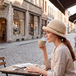 Op de afbeelding: Een vrouw draagt een beige hoed met een bruine band en houdt een beige kopje koffie vast. Ze zit buiten aan een houten tafel, met een krant en een smartphone. De achtergrond toont een Europese straat.