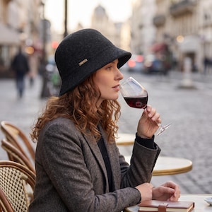 Peut inclure: Une femme portant un chapeau cloche gris foncé avec un petit détail doré, un blazer marron, et buvant du vin rouge dans un verre transparent. La scène se déroule dans un café en plein air avec une rue floue en arrière-plan.