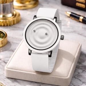 May include: A white wristwatch with a distinctive dial. The watch has a white strap and a circular face with a textured, concentric pattern. It features black accents and a minimalist design. The watch is displayed on a beige stand.