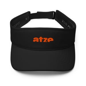 Könnte beinhalten: Ein schwarzes Visier mit dem in orangefarbenem Garn gestickten Wort atze auf der Vorderseite. Das Visier verfügt über einen verstellbaren Riemen mit schwarzer Schnalle auf der Rückseite und das Innenbranding Flexfit 110 am Stirnband.