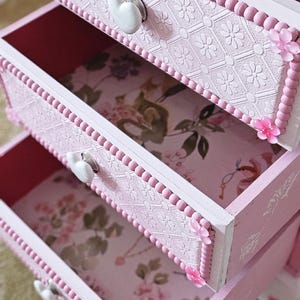 May include: A pink wooden chest of drawers featuring three pull-out drawers with white heart-shaped knobs. Each drawer front is decorated with a textured floral pattern, beaded trim, and small pink flower accents. The interior is lined with floral paper.