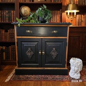 Op de afbeelding: Een zwart en houtkleurig kabinet met een lade en twee deuren. De kast heeft sierlijke hardware in bronskleur en decoratieve accenten. Een witte buste-planter en een wereldbol zijn zichtbaar op de achtergrond. De kast staat op een patroonkleed.