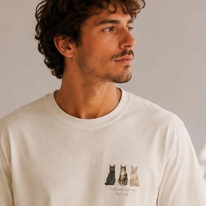 Könnte beinhalten: Ein cremefarbenes Kurzarm-T-Shirt mit einer kleinen Grafik auf der linken Brustseite, die drei Katzen in den Farben Schwarz, Braun und Beige zeigt, mit dem Text "Different colors, Still cat" in kleinen schwarzen Buchstaben darunter.