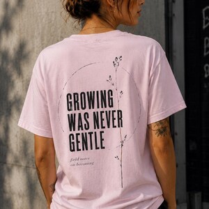 Könnte beinhalten: Ein hellrosa T-Shirt mit kurzen Ärmeln und schwarzem Text auf der Rückseite, der "GROWING WAS NEVER GENTLE" und "field notes on becoming" lautet, neben einer dünnen schwarzen Strichzeichnung eines Pflanzenstängels in einem Kreis.