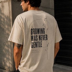Könnte beinhalten: Cremefarbenes T-Shirt mit dem Text "GROWING WAS NEVER GENTLE" in einem Kreis, begleitet von einer botanischen Illustration. Der Satz "field notes on becoming" ist darunter gedruckt. Das T-Shirt wird von einer Person getragen.