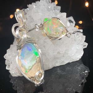May include: Two silver wire wrapped opal pendants. The opals are a milky white with flashes of iridescent color. The pendants are displayed on a bed of white crystal clusters.