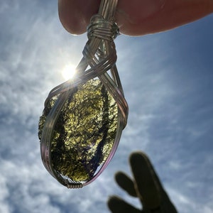 May include: A wire-wrapped pendant featuring a translucent, olive-green, textured stone held in a silver-colored metal frame. The stone has an irregular, jagged surface and is backlit by the sun, highlighting its deep green color.