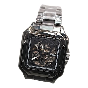 Könnte beinhalten: Schwarze Fossil-Uhr mit quadratischem Zifferblatt und silberfarbenem Metallarmband. Das Zifferblatt hat ein Skelett-Design, das das Innenleben zeigt. Der Markenname "Fossil" ist auf dem Zifferblatt aufgedruckt.
