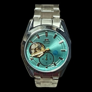 Peut inclure: Une montre automatique Orient argentée avec un cadran turquoise et un mécanisme interne visible. La montre est dotée d'un bracelet argenté et d'un petit cadran en bas. La montre a des accents dorés et est sur fond noir.