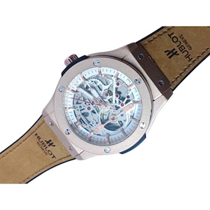 May include: A rose gold Hublot Geneve skeleton watch with a visible mechanical movement, rose gold hands, and a tan leather strap featuring the Hublot Geneve logo. The round case has six visible screws on the bezel and a crown on the side.