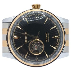 May include: A Seiko Presage Automatic watch with a black sunray dial, gold-tone hands and hour markers, and an open heart aperture. The watch features a gold-tone bezel and a two-tone silver and gold-tone metal link bracelet.