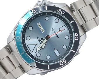 May include: A stainless steel Seiko automatic wristwatch featuring a light blue sunray dial, circular hour markers, and a day-date window at the three o'clock position. The watch includes a rotating bezel with a black and teal insert and a metal link bracelet.