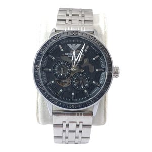 May include: A silver-tone Emporio Armani mechanical wristwatch with a stainless steel link bracelet. The watch features a black skeleton dial with silver hands, indices, and two sub-dials. The Emporio Armani logo is positioned at the twelve o'clock mark.