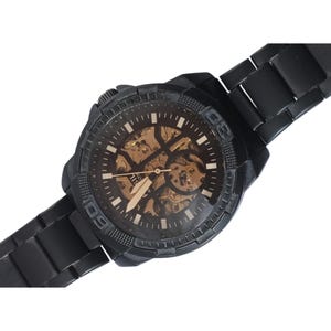 May include: A black wristwatch with a metal link band and a round case. The dial features a skeletonized design revealing gold-toned internal mechanical components, white hour markers, and luminous hands. The bezel displays engraved numerals.