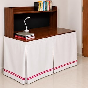 May include: A wooden desk with a hutch, featuring a white fabric skirt with two horizontal pink stripes along the bottom edge. The desk surface holds a white desk lamp and a stack of books, while the hutch shelf contains a row of upright books.