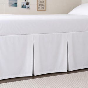 Könnte beinhalten: Ein weißer Bettrand mit plissiertem Design wird auf einem Bett mit einem weißen Spannbetttuch und Kissen gezeigt. Der Bettrahmen ist aus Holz. An der Wand hinter dem Bett befinden sich kleine gerahmte Bilder und ein Schild mit der Aufschrift "FOCUS WORK BE KIND".
