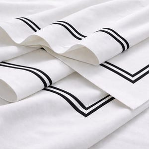 May include: White bed sheets with black trim. The sheets are folded and draped, showcasing the contrasting black lines that frame the edges and create a modern, minimalist aesthetic. The bedding is crisp and clean.