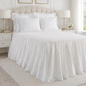 May include: A white bed with a ruffled bed skirt, pillows, and a tufted headboard. Two framed botanical prints hang above the bed. Two lamps sit on bedside tables. The room has a neutral color palette.