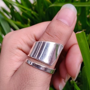 May include: A wide, polished silver-tone ring featuring a dual-band design. The upper portion is a broad, curved plate with a smooth, reflective surface, while the lower portion consists of a thinner, plain band that wraps around the finger.