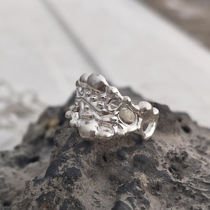 May include: A silver ring with an organic, textured design. The ring features a bubbly, irregular surface with small, rounded protrusions. The ring is set on a dark, textured rock, with a blurred white background.